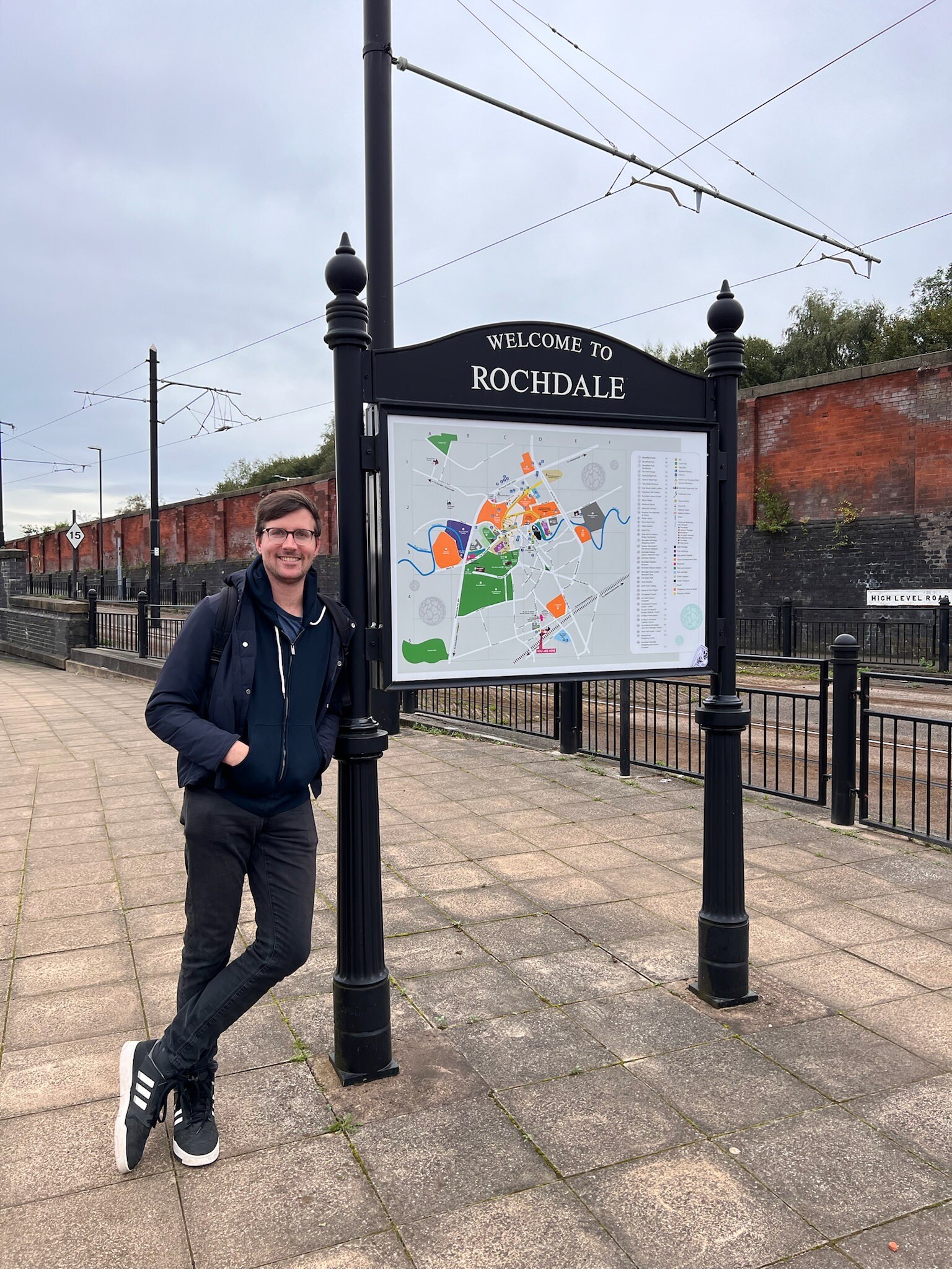 A smiling dude standing next to a sign that says “Welcome to Rochdale” and displays a town map