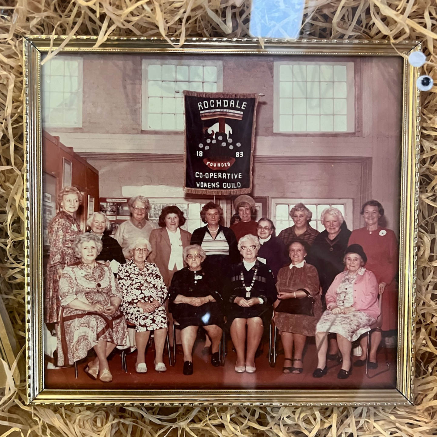 A historical photo of the Rochdale Cooperative Women’s Guild, with several women sitting under a banner in a half-circle, looking at the camera