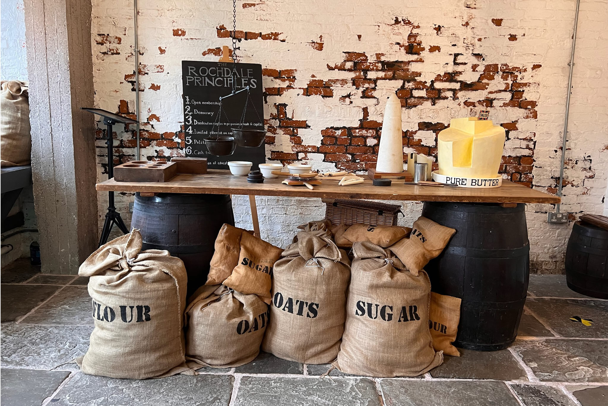 An improvised table with a wooden plank over two barrels, butter on top, sacks of flour, sugar and oats on the floor. It’s a scene from the Rochdale Pioneers Museum, mimicking the appearance of the first Pioneers store in 1844 at Toad Lane, Rochdale.