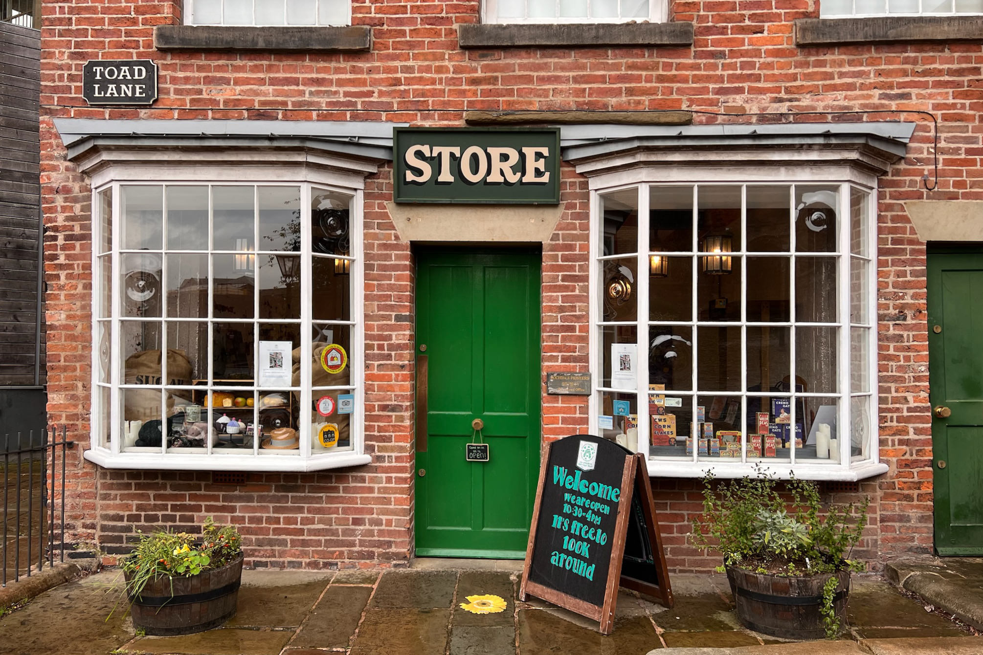 A quaint English storefront with a sign that says “Store” and two large windows, a green door, in a red brick building