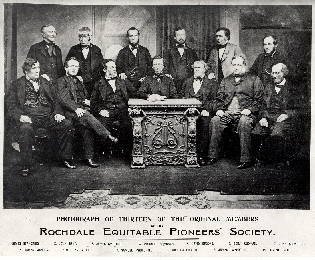 A historical photo of 13 of the original Rochdale pioneers. Lots of men in suits sitting in a half circle, serious atmosphere, with the name of their company written below: Rochdale Equitable Pioneers’ Society