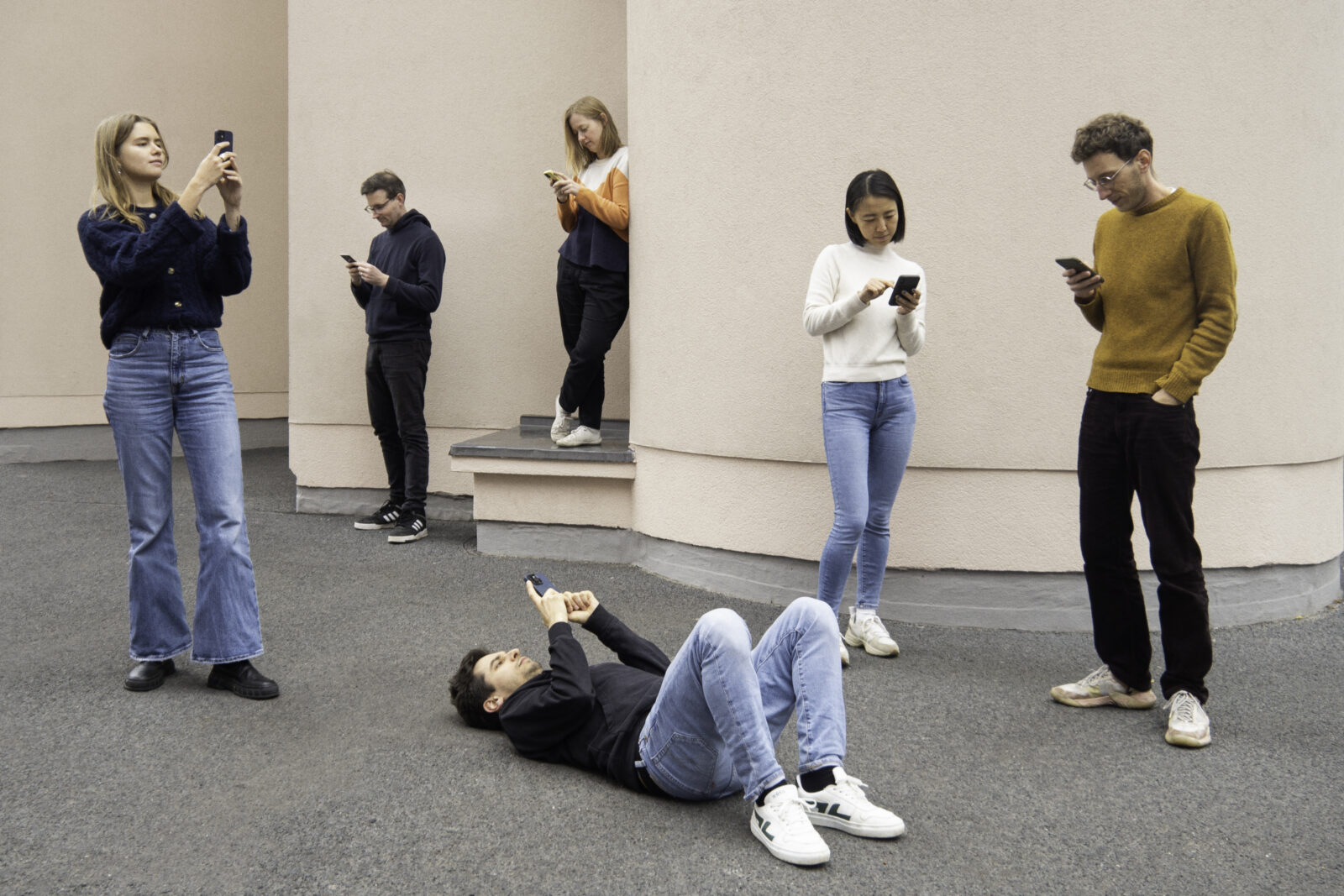 A team photo of all our six members, standing in front of a building wall, all looking at their phones, one person lying on the floor