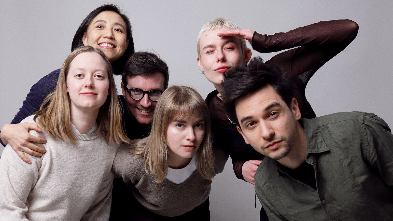 Team photo of the entire Village One team, with six people standing close together, looking at the camera, some laughing, some looking skeptical, some hugging. Relaxed mood in a professional photo studio setting, with a light beige background.