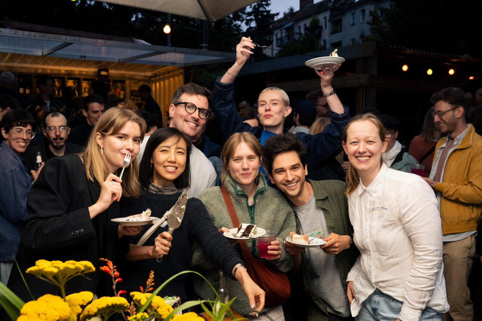 Photo of an anniversary party, outside in a beer garden, at dusk, a group of people standing close together, eating cake and smiling at the camera