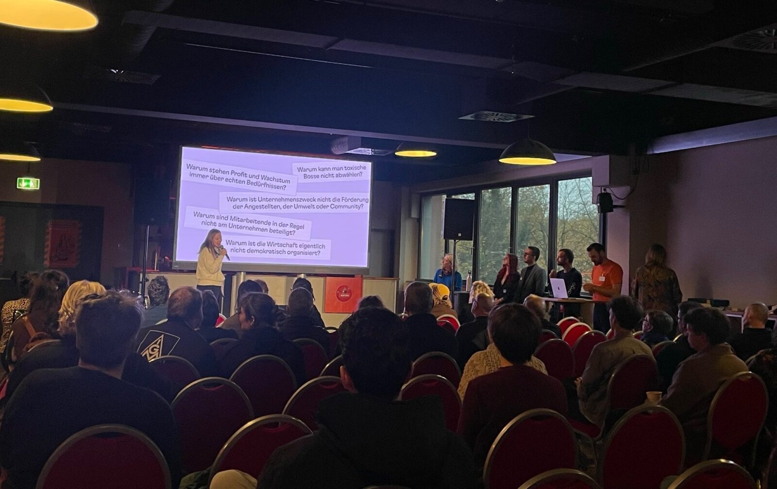 Photo of a packed conference room, with a large projector, a woman on stage with a slide behind her asking questions like “Why can’t you un-elect toxic bosses?” or “Why is there so little democracy in the economy?”