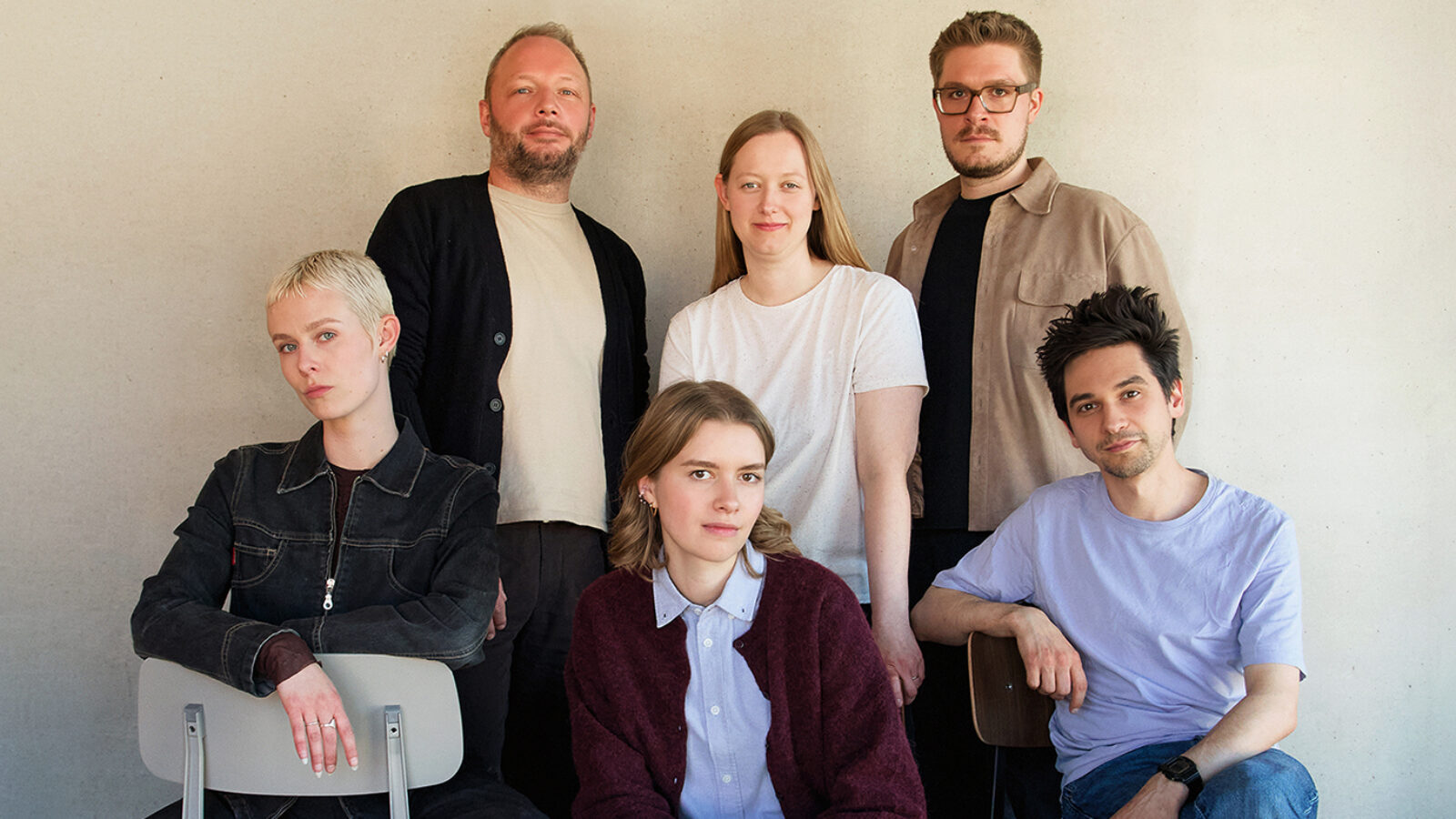 Photo of the Village One team, showing Meg, John, Julia, Doro, Niklas and Harry (from left to right), sitting and standing close together in front of a concrete wall, all looking at the camera, some of them slightly smiling.