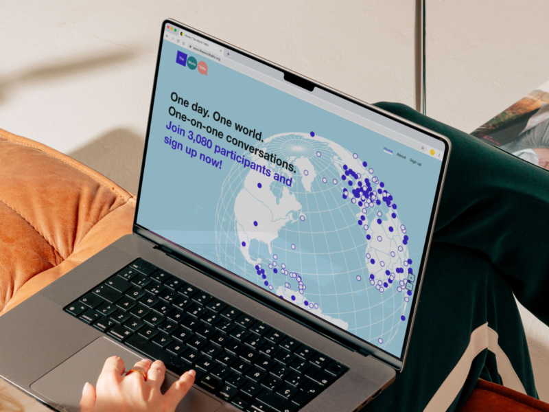 Over-the-shoulder photo of a person sitting on a couch with a laptop. The screen shows a website advertising a world-wide event in which different people can meet. It’s visualized with an abstracted globe with dots for every country.
