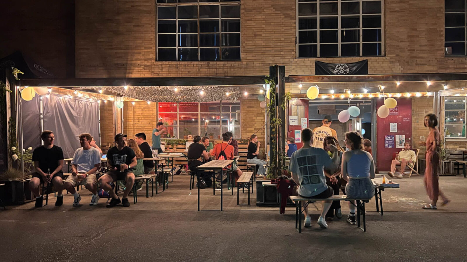Photo of the outside area of a bar, with approximately 20 people sitting and walking around in a casual setting, at night
