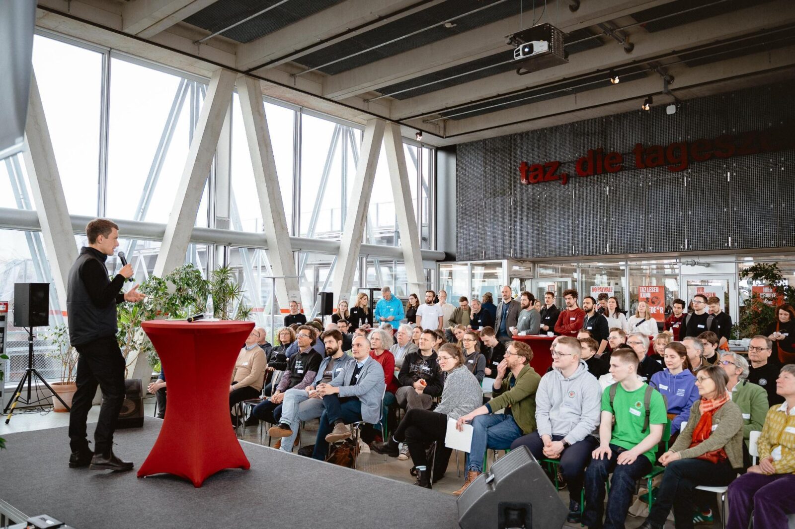 A photo from the stage of a conference, with a male speaker on stage holding a microphone and a large audience listening, in a modern building with exposed concrete beams