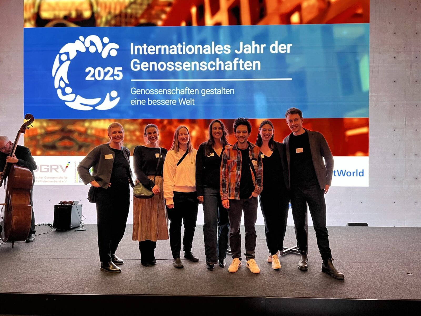 A photo of a group of people standing on a stage, posing for the camera, with a projection in the background that says “2025: Internationales Jahr der Genossenschaften”