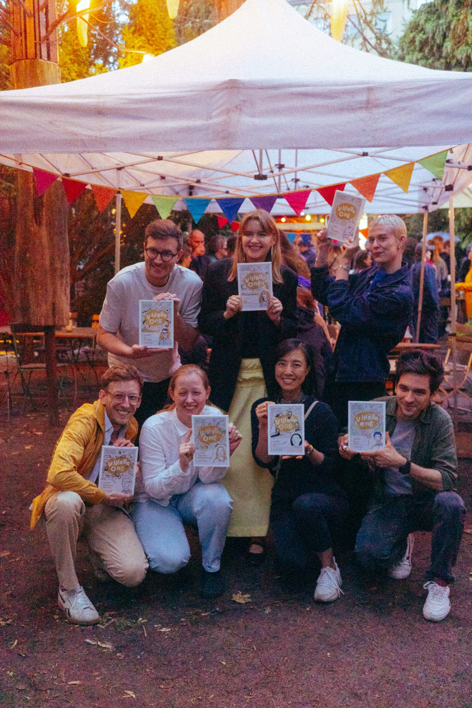 Photo of the entire Village One team, standing and kneeling close together as a group, smiling, holding up framed membership certficates, outside setting with a gazebo, lots of people visible in the far background, warm lighting