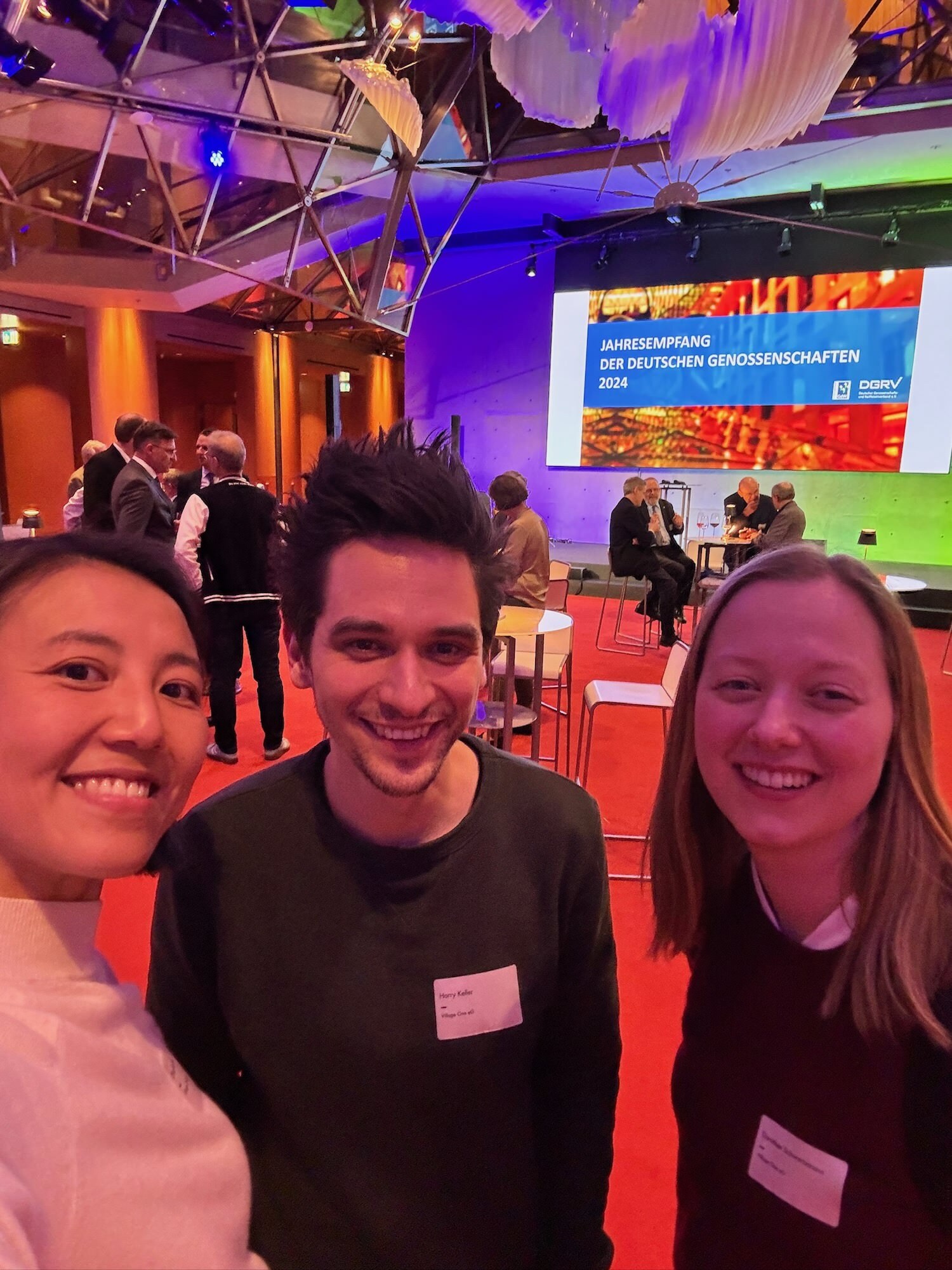 Selfie of three people from Village One in an event space, stage visible in the background with a powerpoint slide that says “Jahresempfang der deutschen Genossenschaften”, colorful lighting