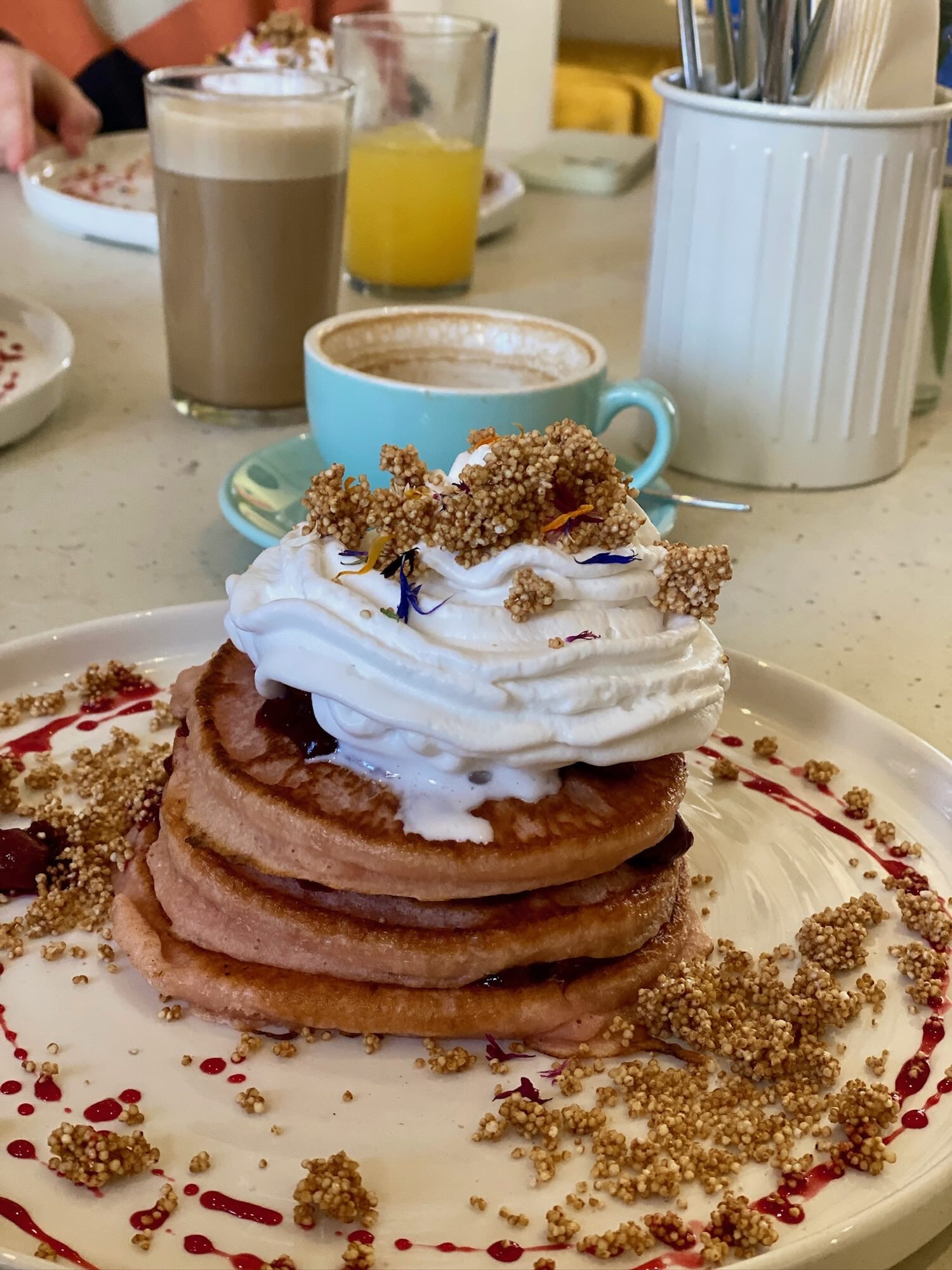 A photo of a large pile of vegan pancakes, with coconut cream on top, coffee cups in the background