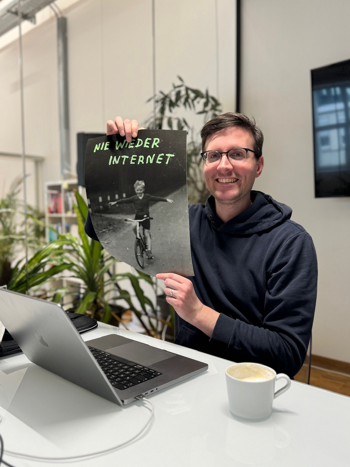 One person in a meeting room, smiling and holding a poster that shows a child riding a bicycle with the handwritten caption “Nie wieder Internet”, emphasizing the joy of an analog lifestyle